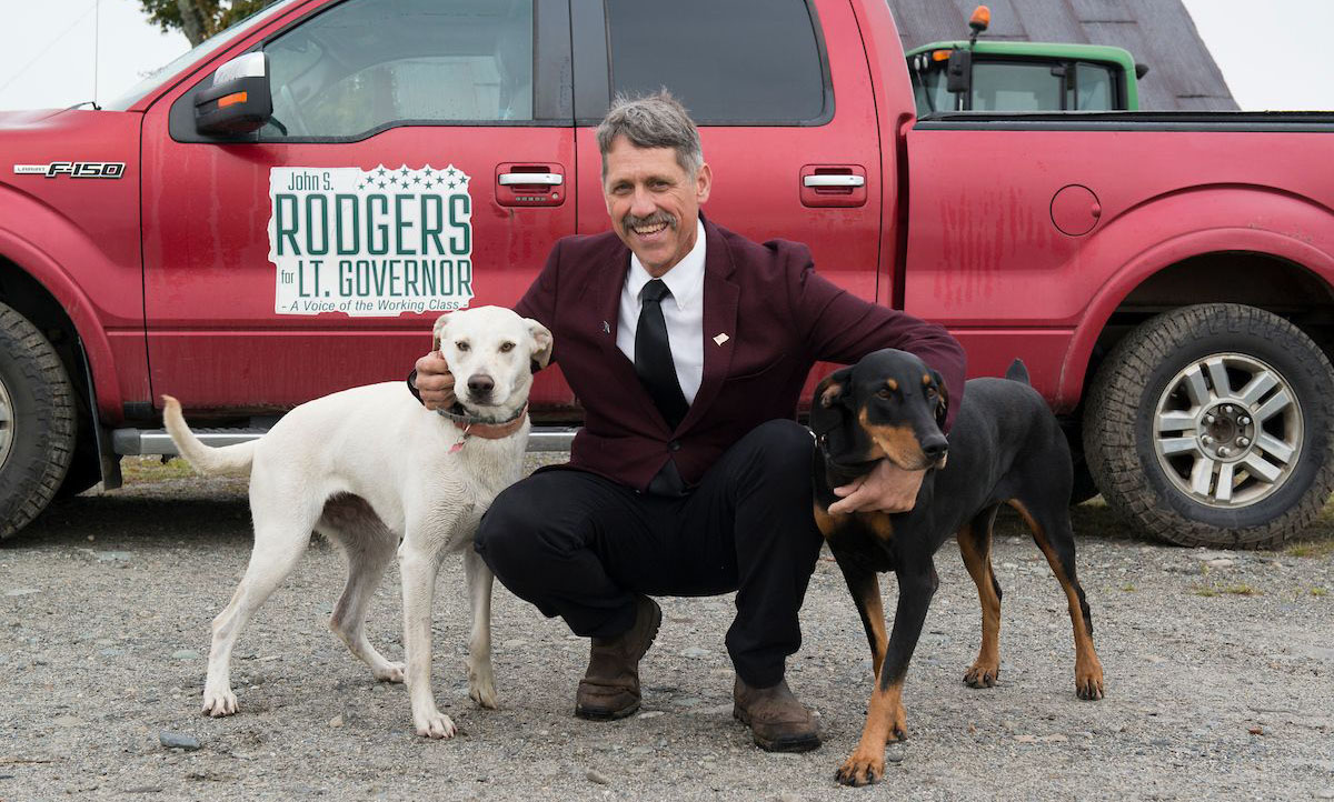 Lieutenant Governor John Rodgers is crouching and smiling, wearing a burgundy blazer, black tie, and black pants. He is flanked by two dogs—a white dog on his left and a black-and-tan dog on his right. Behind him is a red pickup truck with a campaign sign that reads, "John S. Rodgers for Lt. Governor - A Voice of the Working Class." The setting appears to be outdoors with a gravel ground and farm equipment in the background.